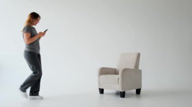 Beyaz arka planda cep telefonuyla koltukta dinlenen neşeli beyaz bir kadın. Dijital eğlence ve online eğlence kavramı.