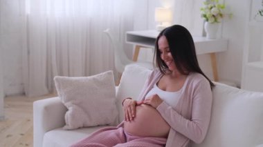 Pozitif orta yaşlı beyaz kadın hamile karnına hafifçe dokunuyor, evdeki kanepede dinleniyor. Kadınlık ve bebek beklentisi kavramı.