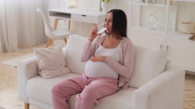 Hamile, orta yaşlı, beyaz, pozitif kadın evde camdan su içiyor. Sağlıklı hamilelik, susuzluk ve ferahlama kavramı.