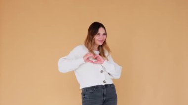 Mutlu ve duygusal Kafkasyalı genç bir kadın, bej arka planda duran kalp şeklindeki işaretle alay ediyor ya da flört ediyor. Aşk ya da jest gibi.