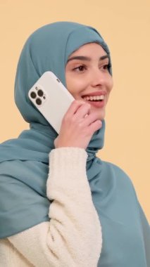 Başörtüsü takan mutlu Arap kadın bej arka plandaki telefon görüşmesiyle sohbete daldı. Uzaktan iletişim konsepti. Dikey video.