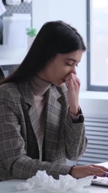 Ofiste çalışırken burnu akan, peçeteye hapşıran, hasta, genç, beyaz bir kadın. Soğuk algınlığı ve grip kavramı. Dikey video.