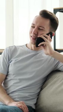 Mutlu, genç, beyaz bir adam cep telefonu kullanıyor, arkadaşlarıyla konuşuyor ve ararken gülüyor, evde boş zaman geçiriyor. Dikey video.