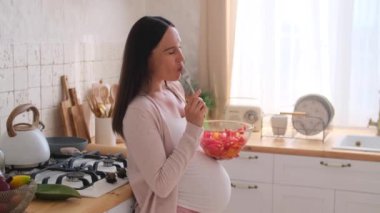 Heyecanlı, orta yaşlı, hamile bir kadın. Kaseden sağlıklı salata yiyor, ev mutfağında duruyor. Hamilelik emekliliği kavramı.