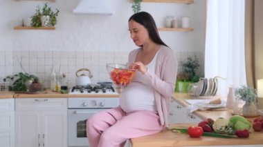 Orta yaşlı mutlu bir beyaz kadın sebze salatasından zevk alıyor, evdeki mutfak masasında oturuyor. Taze ve sağlıklı yemek konsepti.