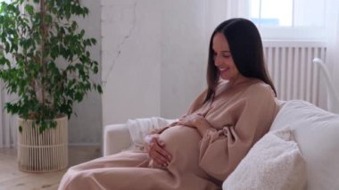 Mutlu, orta yaşlı, beyaz bir kadın oturma odasındaki kanepeye uzanmış bebek yumrusunu okşuyor. Gelecekteki ebeveyn kavramı.