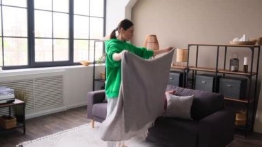 Evdeki oturma odasında yastıklı ve battaniyeli kanepeyi düzenleyen orta yaşlı neşeli beyaz kadın ya da ev hanımı. Günlük günlük ev işleri kavramı.