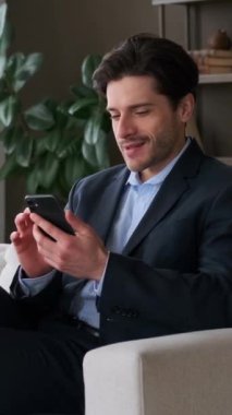 Gülümseyen beyaz erkek çalışan online eğlence için akıllı telefonu kullanıyor, boş zamanlarını evde koltukta dinlenerek geçiriyor. Dikey video.