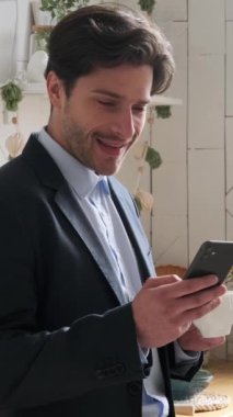Akıllı telefon kullanan, internet üzerinden haber okuyan, sabah rutininde sıcak içecek içen neşeli beyaz erkek çalışan. Dikey video.