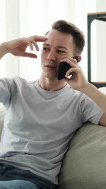 Güler yüzlü, genç, beyaz bir adam akıllı telefonu uzaktan iletişim için kullanıyor, boş zaman geçiriyor ve evde gülüyor. Dikey video.