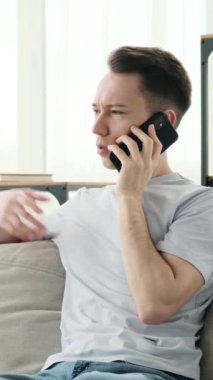 Ciddi bir Kafkasyalı genç adam akıllı telefon kullanarak sohbet ve pazarlıkla meşgul, evdeki kanepede oturuyor. Uzaktan iletişim kavramı. Dikey video.