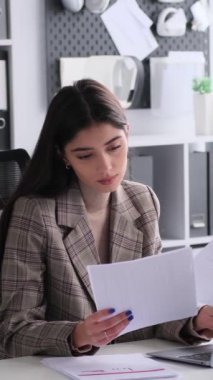 Düşünceli, beyaz, genç, serbest çalışan bir kadın büroda dizüstü bilgisayarla çalışıyor ve ifadeleri analiz ediyor. Dikey video.