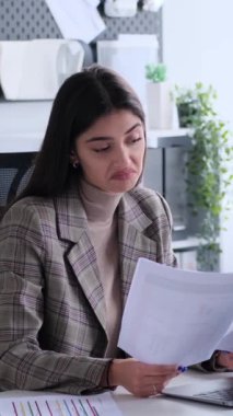 Kafası karışmış, genç, beyaz, yetişkin bir iş kadını büroda evraklarla çalışırken kollarını açıyor. İkilem kavramı. Dikey video.
