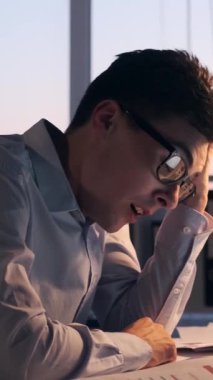 Uykucu, genç, beyaz bir adam akşam çalışırken ofiste dizüstü bilgisayarla esniyor. Yorgunluk ve can sıkıntısı kavramı. Dikey video.