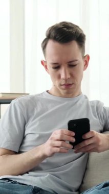Konsantre olmuş genç beyaz adam evdeki kanepede dinlenirken cep telefonuyla çevrimiçi sosyal medyada geziniyor. Dikey video.