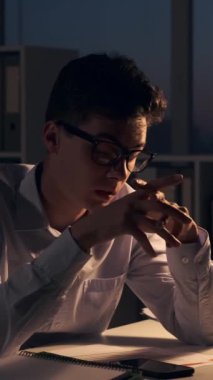 Yorgun, genç, beyaz, serbest çalışan bir erkek. Akşam ofisinde çalışırken cep telefonu kullanıyor. Dikey video.
