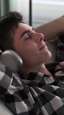 Hafta sonu boş vakitlerinde kanepede dinlenirken müzik dinleyen heyecanlı beyaz bir genç adam. Dikey video.