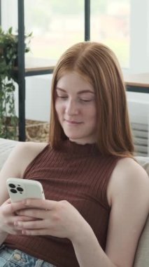 Kızıl saçlı, mutlu, genç bir kız telefonunu online eğlence ve mesajlaşma için kullanıyor, boş zamanlarını evde geçiriyor. Dikey video.