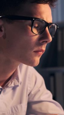 Gece geç saatlerde ofiste dizüstü bilgisayarla ve belgelerle çalışan odaklanmış genç bir beyaz erkek çalışanın portresi. Dikey video.