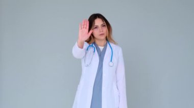 Mutsuz ve ciddi bir beyaz kadın doktor ya da hemşire dur işareti gösteriyor ve gri arka planda başını sallıyor. Uyarı ya da reddetme hareketi.
