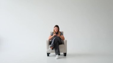 Beyaz arka planda cep telefonu kullanarak internette gezinen, gülen beyaz, genç bir kadın. Çevrimiçi sosyal medyada mesajlaşmak, dijital zaman harcamak.