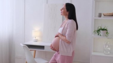 Neşeli, orta yaşlı, beyaz bir kadın. Evdeki bebek şişliğini şefkatle ovalıyor. Gelecekteki anneliğin rahatlığı ve keyfi..