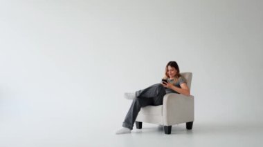 Kafkasyalı genç kadın internette geziniyor ve beyaz arka planda telefonuyla koltukta dinleniyor. Siber uzayda eğlence ve keyif.