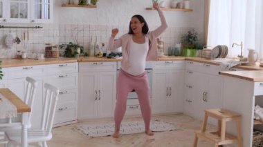 Orta yaşlı hamile, beyaz, dost canlısı kadın ev mutfağında müzik eşliğinde dans ediyor. Aktif eğlence ve mutlu hamilelik kavramı.