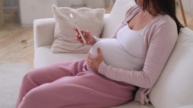 Hamile, orta yaşlı, beyaz bir kadının sosyal medyada cep telefonuyla sohbet ederken ve karnına dokunurken yakın plan çekimleri..