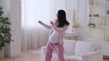 Pozitif beyaz kadın hamilelikten zevk alıyor, müzik eşliğinde dans ediyor, zevk alıyor ve rahat bir evde oturma odasında eğleniyor..