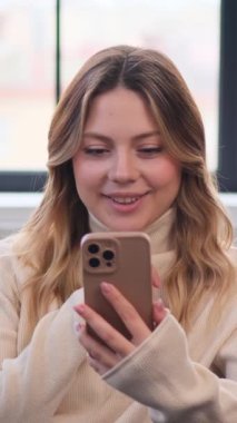 İnternette cep telefonu kullanan ve evde boş zamanlarında gülen mutlu, genç beyaz bir kadın. Dikey video.