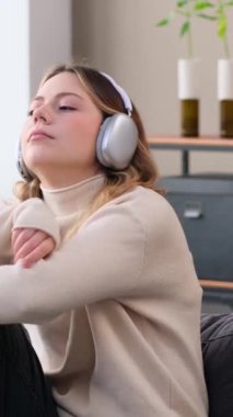 Kederli, genç, beyaz bir kadın kulaklıkla müzik dinliyor ve boş zamanlarını evde geçiriyor. Depresyon kavramı. Dikey video.