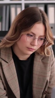 Mutsuz ve yorgun, genç, beyaz bir kadın ofiste masa başında çalışıyor. Üzüntü, yorgunluk ve hüsran konsepti. Dikey video.