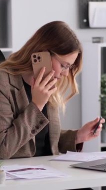 Ofiste belgelerle çalışan ve telefon görüşmesiyle meşgul olan konsantre olmuş Kafkasyalı genç kadın. Sohbet ya da pazarlık kavramı. Dikey video.