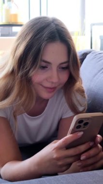 Keyifli bir kadın boş zamanlarını telefonuyla, internet eğlencesiyle ya da internet sohbetleriyle geçiriyor, evdeki kanepede uzanıyor. Dikey video.