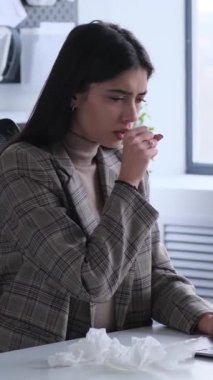 Hasta, genç, beyaz bir iş kadını, hastalık ve burun akıntısından muzdarip, modern ofiste çalışırken öksürüyor. Dikey video.