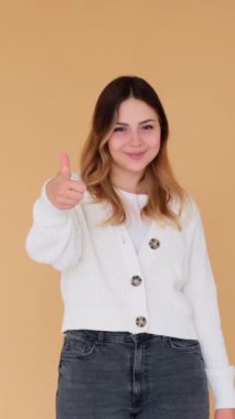 Gülümseyen Kafkasyalı genç bir kadın tatmin edici bir jest yapıyor ve bej arka plan konusunda hemfikir. Destek işareti. Dikey video.