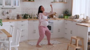Güler yüzlü, orta yaşlı, beyaz, hamile bir kadın ev mutfağında müzik eşliğinde dans ediyor. Mutlu bir hamilelik ve iyi bir ruh hali kavramı.