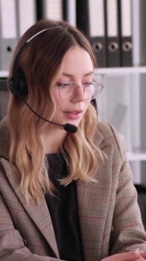 Ofiste çalışırken kulaklık kullanarak müşteriyle konuşan neşeli, beyaz, genç bir kadın müşteri destek ajanı. Dikey video.