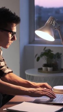 Bilgisayarla çalışan ya da internetten öğrenen konsantre olmuş genç beyaz bir adam, akşam oturma odasında masa başında oturuyor. Dikey video.
