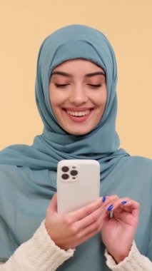 Mutlu Arap genç kadın cep telefonunu sosyal medyada sohbet etmek ve bej arka planda gülmek için kullanıyor. Dikey video.