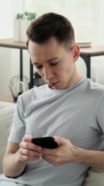 Kanepede dinlenen ve evde online eğlence için telefon kullanan ciddi bir Kafkas genci. İnternet iletişimi kavramı. Dikey video.