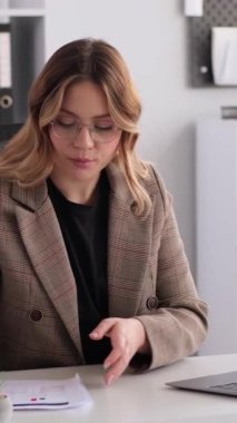 Ciddi, genç, yetişkin, beyaz kadın ofis çalışanı büroda evrak işleriyle uğraşıyor ve video görüşmeleri yapıyor. Çevrimiçi konferans ve web sitesi kavramı. Dikey video.