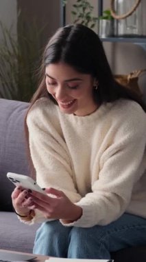 Mutlu, genç, beyaz bir kadın cep telefonu kullanıyormuş. Evde boş vakitlerini geçiriyormuş. Sosyal medyayla iletişim kuruyormuş. Dikey video.