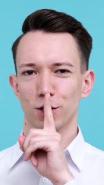 Olumlu, genç, beyaz bir adam sessiz kalma ya da mavi arka planda mahremiyet sağlama konusunda sessiz hareketler sergiliyor. Dikey video.