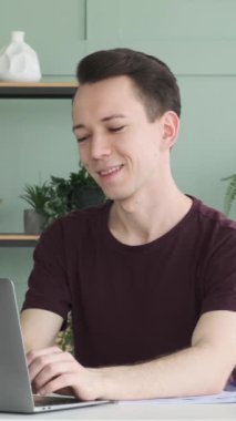 Gülümseyen ve mutlu genç Kafkasyalı erkek ofis bilgisayarını kullanıyor. Uzaktan çalışma ya da öğrenim kavramı. Dikey video.