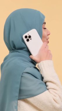 Neşeli ve pozitif Arap genç bir kadının portresi. Konuşurken akıllı telefonu kullanıyor, bej arka planda duruyor. Dikey video.