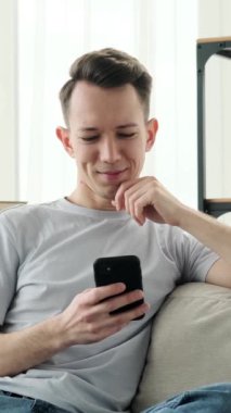 Genç, güler yüzlü, beyaz bir adam rahat evdeki kanepede dinlenirken internetten sosyal medyada telefonla mesajlaşıyor. Dikey video.