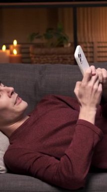 Yorgun, beyaz, genç bir adam kanepeye uzanmış, telefonu internette mesajlaşmak ve gece yarısı esnemek için kullanıyor. Sıkıntı ve yorgunluk kavramı. Dikey video.