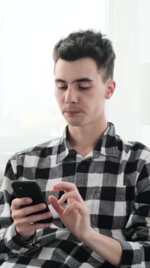 Mutlu, genç, beyaz bir adam internette sohbet ediyor ve gülüyor. Oturma odasında boş vakitlerini telefonda geçiriyor. Dikey video.